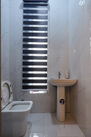 Une salle de bain équipée de toilettes, d’un lavabo et d’une fenêtre avec des stores.