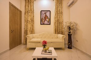 A living room with a beige sofa, a white coffee table with books and a golden lion statue, a framed artwork on the wall, and a tall vase with flowers. - Javes Luxury Residences (Lagos)
