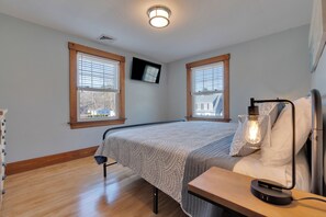 A bedroom with a bed, a bedside table with a lamp, two windows with blinds, and a TV mounted on the wall. - Pet-Friendly York Beach Vacation Rental | Walk to Cape Neddick Beach (York)