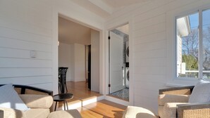 Ein geräumiges Wohnzimmer mit Holzboden, weißen Wandverkleidungen und einem großen Fenster. Es gibt zwei Korbsessel mit Kissen und einen kleinen runden Tisch.