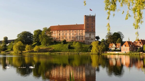 Un edifici històric amb una torre, ubicat al costat d'un llac tranquil amb oques, envoltat de vegetació exuberant i arquitectura tradicional.