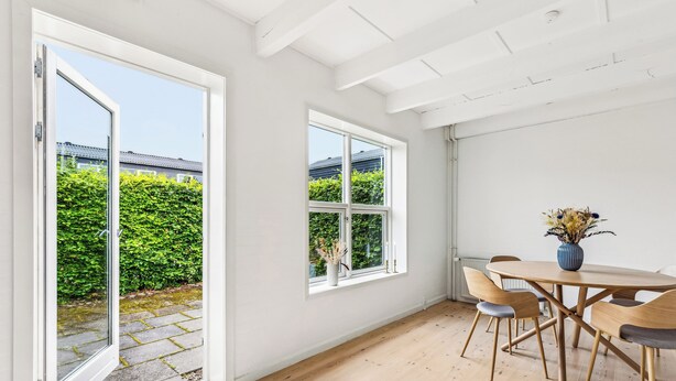 Una habitación con piso de madera, una mesa redonda con sillas, un florero con flores secas y una ventana que ofrece vistas a zonas verdes y edificios.