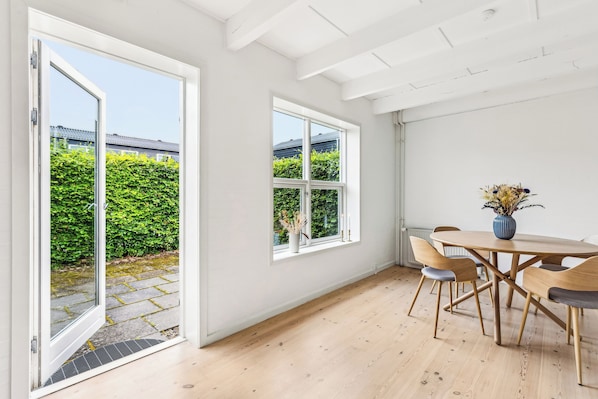 Una habitación con piso de madera, una mesa redonda con sillas, un florero con flores secas y una ventana que ofrece vistas a zonas verdes y edificios.