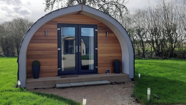 A small, semi-open wooden structure with a dark door and glass panels, surrounded by a grassy area and trees. - Lotus Pod Self-Catering glamping in Suffolk (Henley)