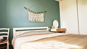 Un dormitorio con cama, una silla, una mesita de noche con espejo y un colgador de macramé en la pared.