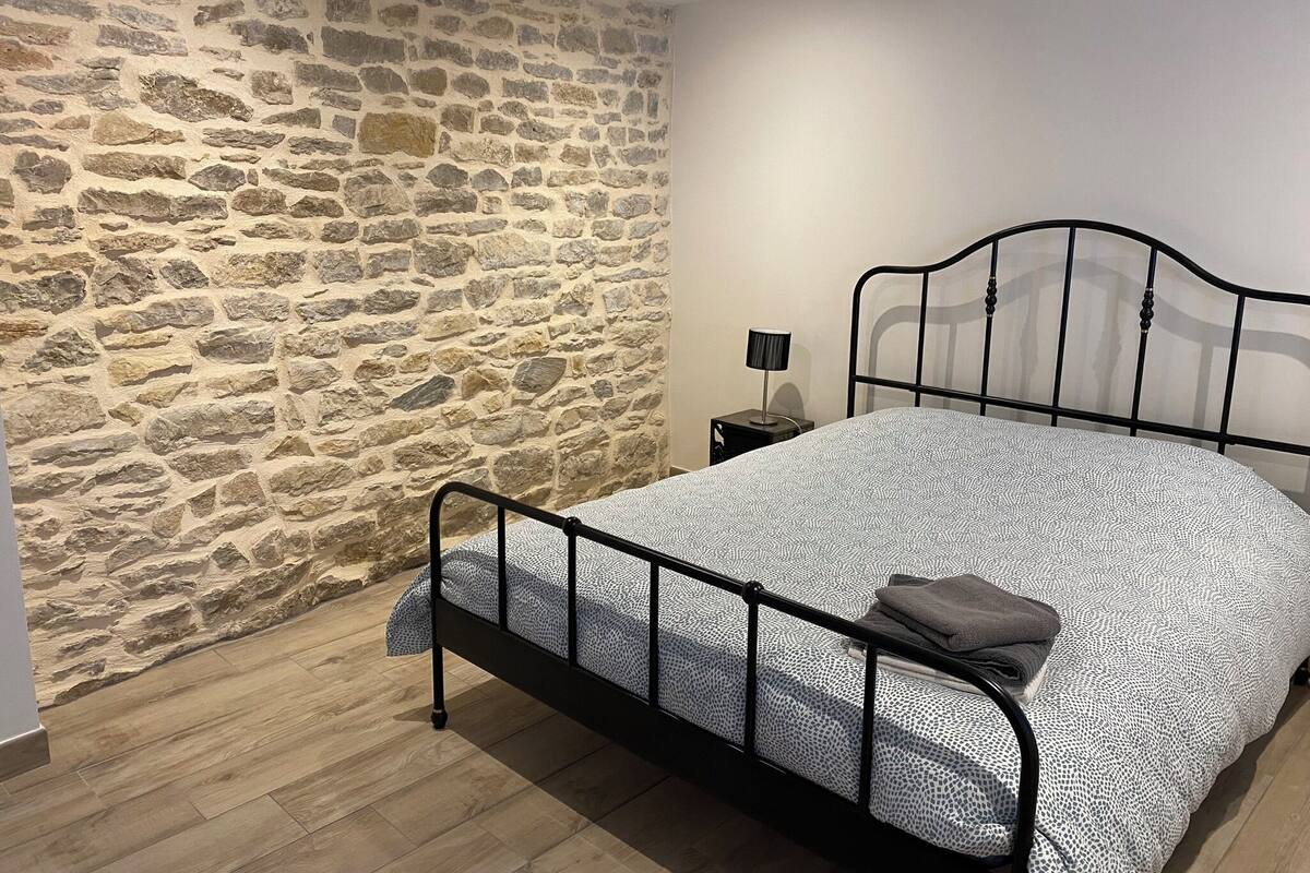 A bedroom with a stone wall, a metal bed frame, a patterned bedspread, and a bedside lamp.
