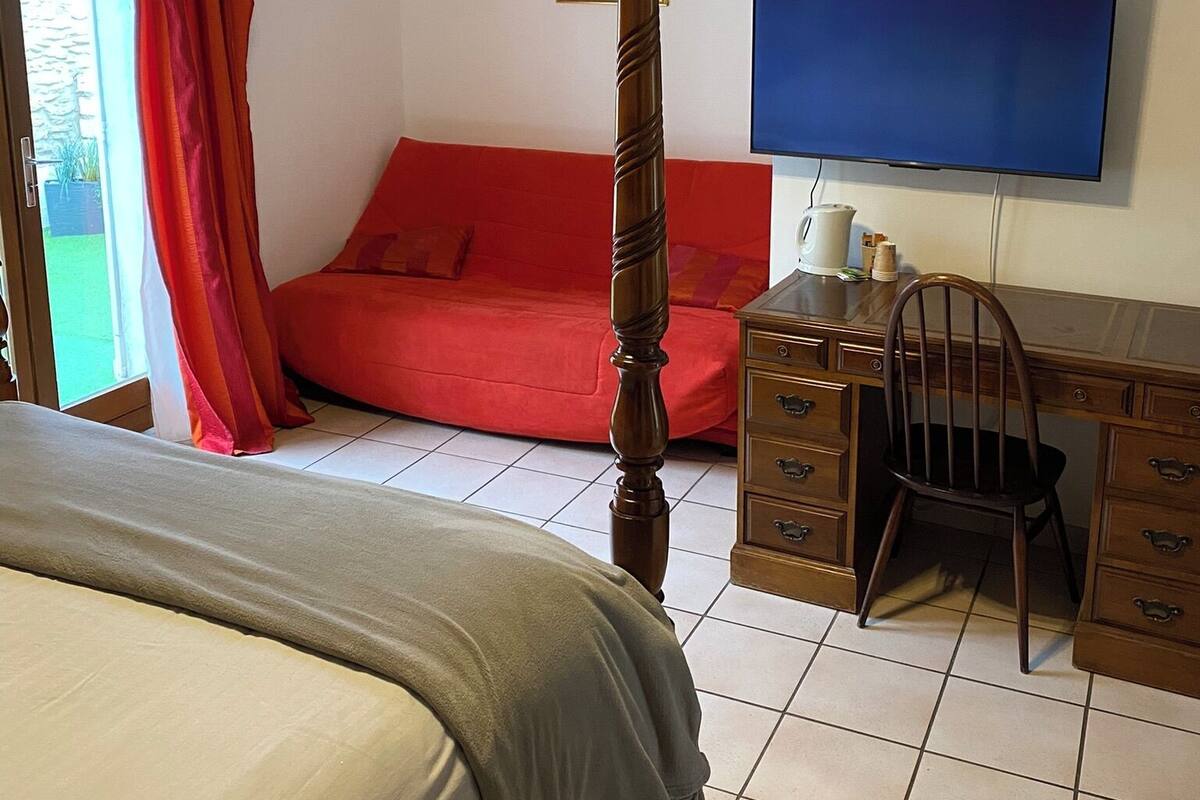 A bedroom with a four-poster bed, a red sofa, a wooden desk with drawers, and a television on the wall.