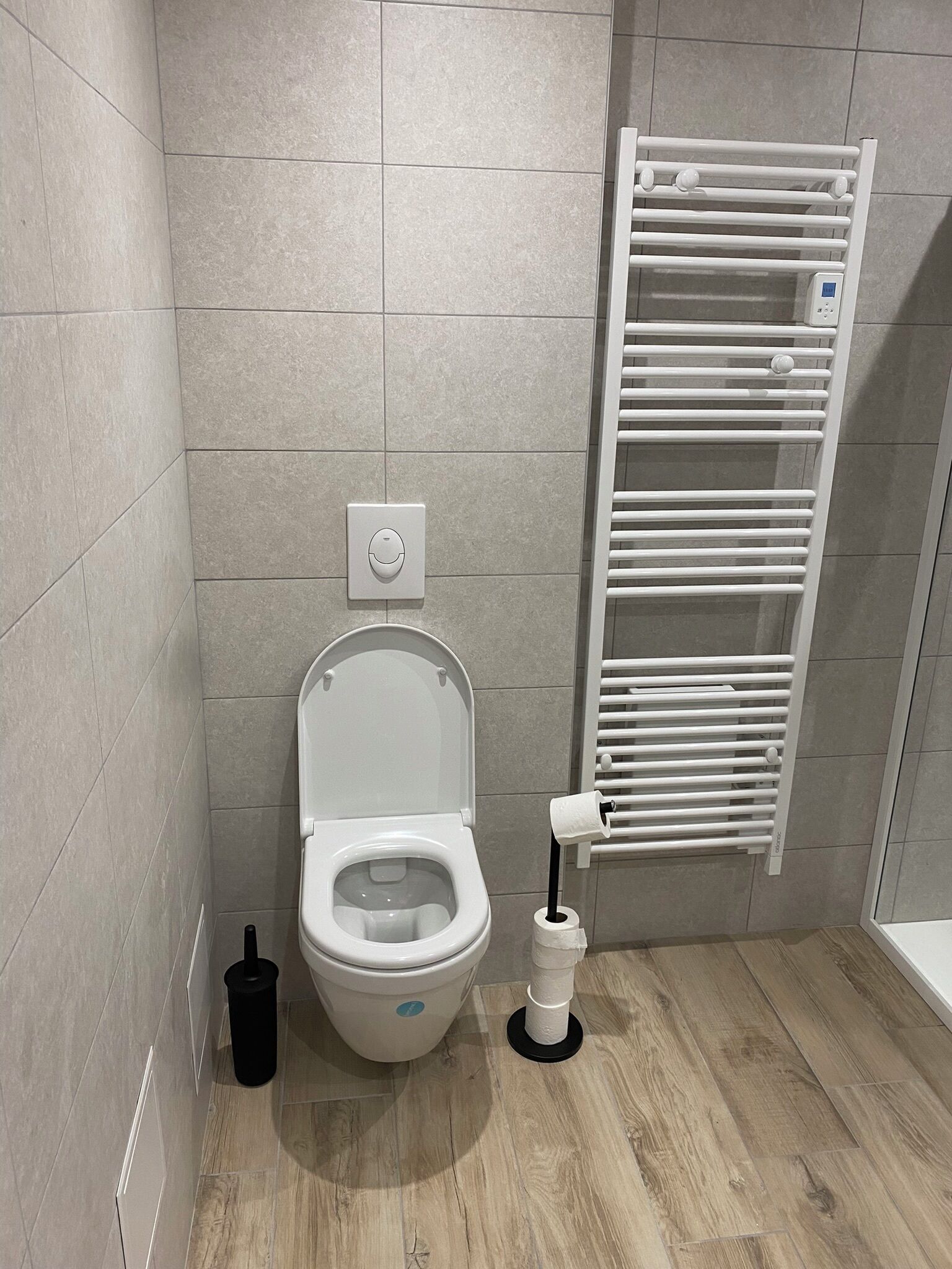 A modern bathroom with a toilet, a wall-mounted flush button, a toilet paper holder, and a white towel rack.