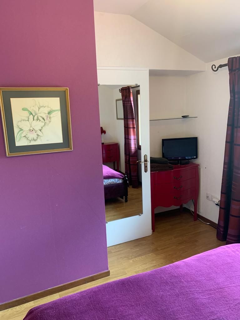 Une chambre avec un mur violet, un lit recouvert d’une couverture violette, une commode rouge, un téléviseur posé sur un meuble et un tableau encadré accroché au mur.
