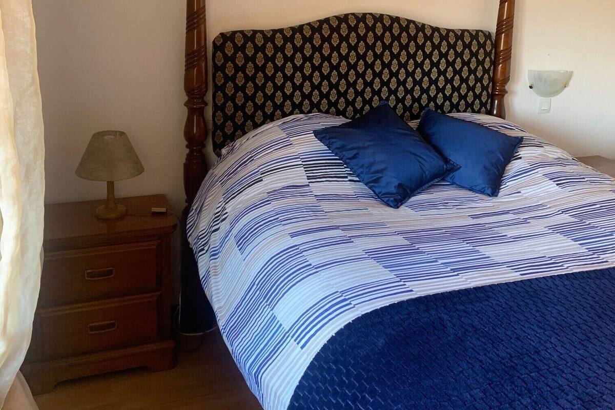 A bedroom with a four-poster bed, a patterned headboard, a framed picture on the wall, a bedside table with a lamp, and a nightstand with drawers.