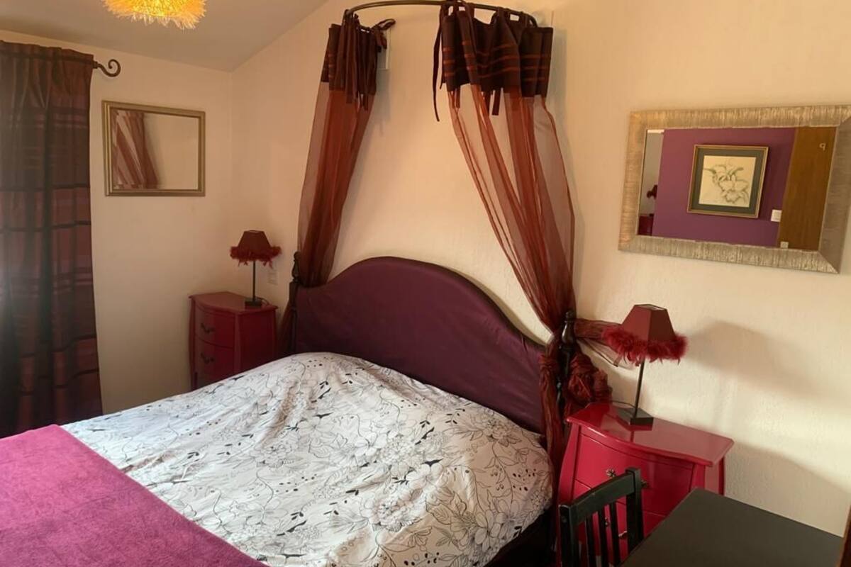 A room with a bed, a nightstand, a chair, a table, a lamp, a mirror, and curtains.