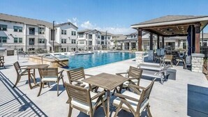 Un espace piscine doté de chaises longues et de tables, entouré d’immeubles à plusieurs étages avec des balcons.