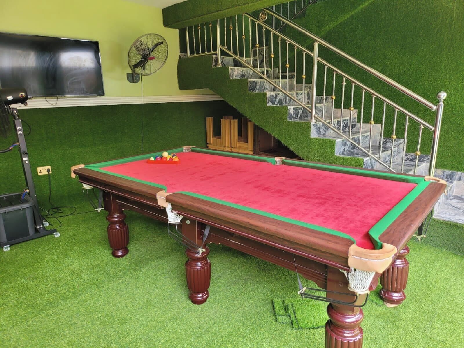 Uma mesa de bilhar com superfície de feltro vermelho, cercada por grama sintética verde.