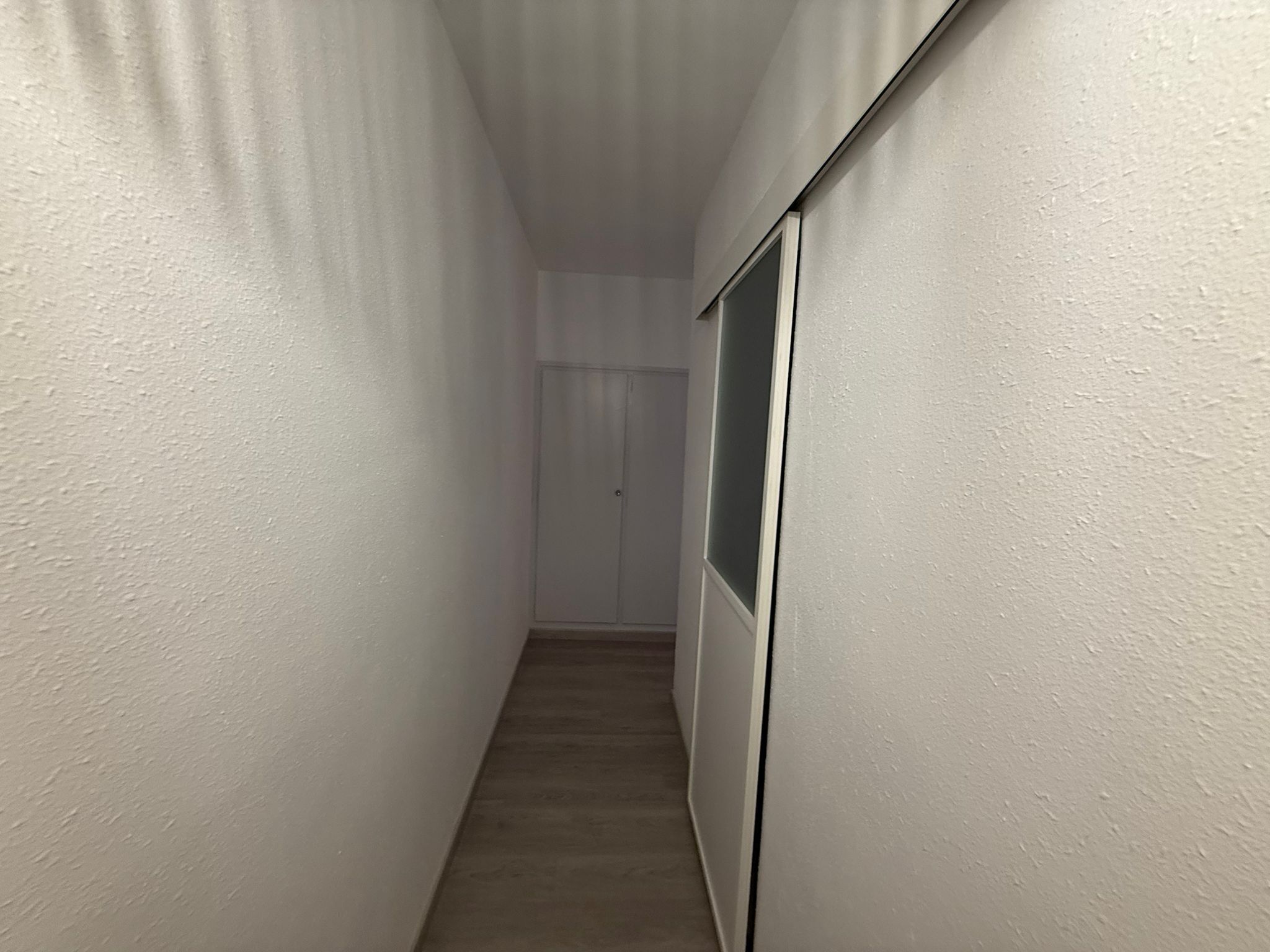 Ein schmaler Gang mit weißen Wänden und Holzboden, der zu einer Tür am Ende führt.