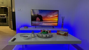 Una sala de estar moderna con un televisor de pantalla plana montado sobre un mueble blanco, una mesa de centro con un control remoto, un tazón y una caja de galletas Bastogne.