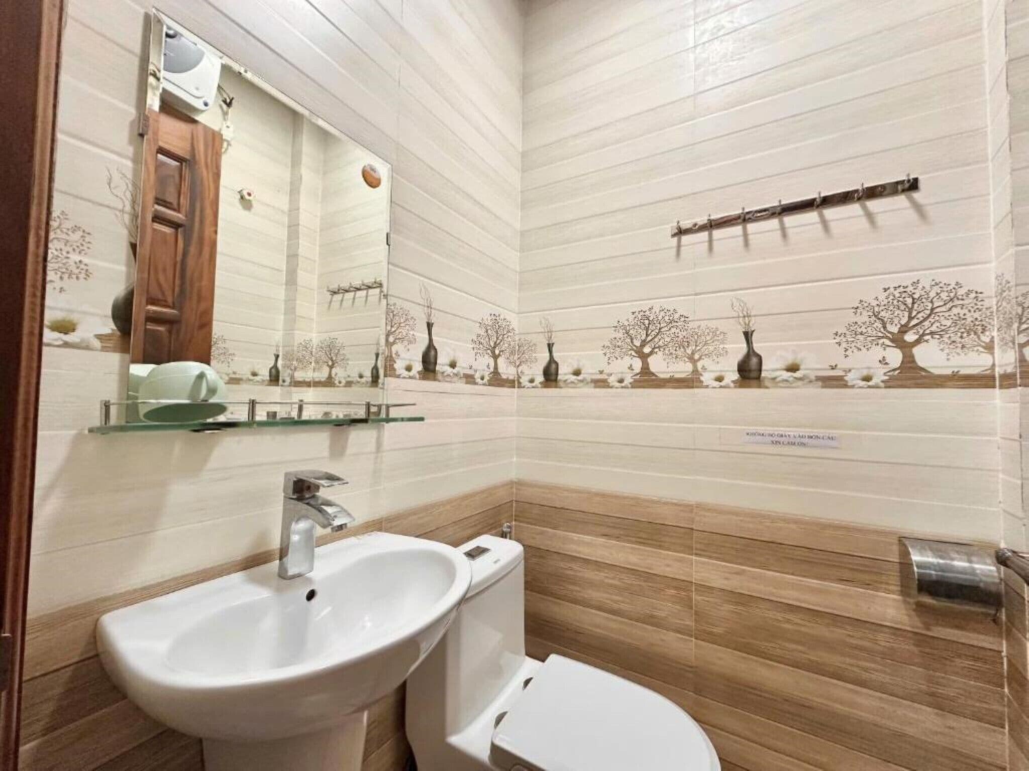 A bathroom with a white sink, a toilet, and a mirror. There are decorative trees on the wall and a shelf with a vase.