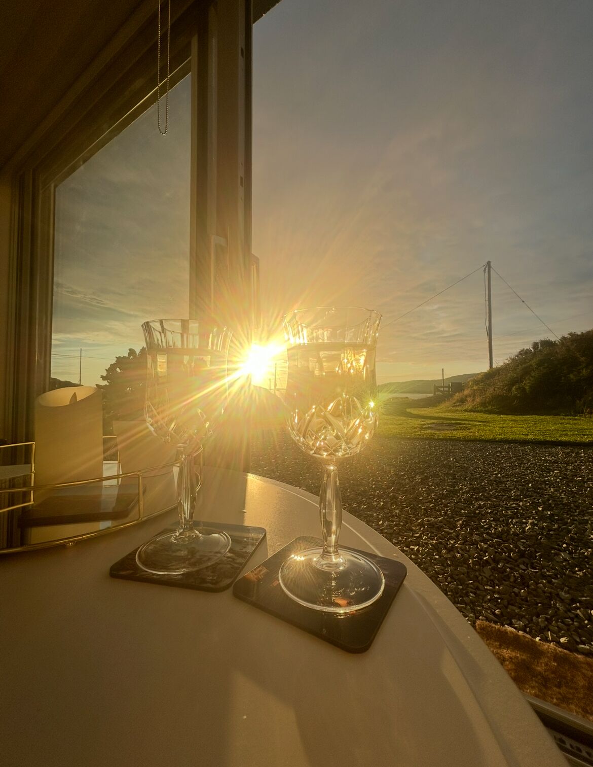 A table with two glasses of water and a decorative glass centerpiece, viewed through a window overlooking a gravel path and greenery.