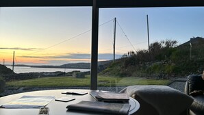 Een tafel met een afstandsbediening, een tijdschrift en een boek, met uitzicht op een kustlandschap met heuvels en een zonsondergang.