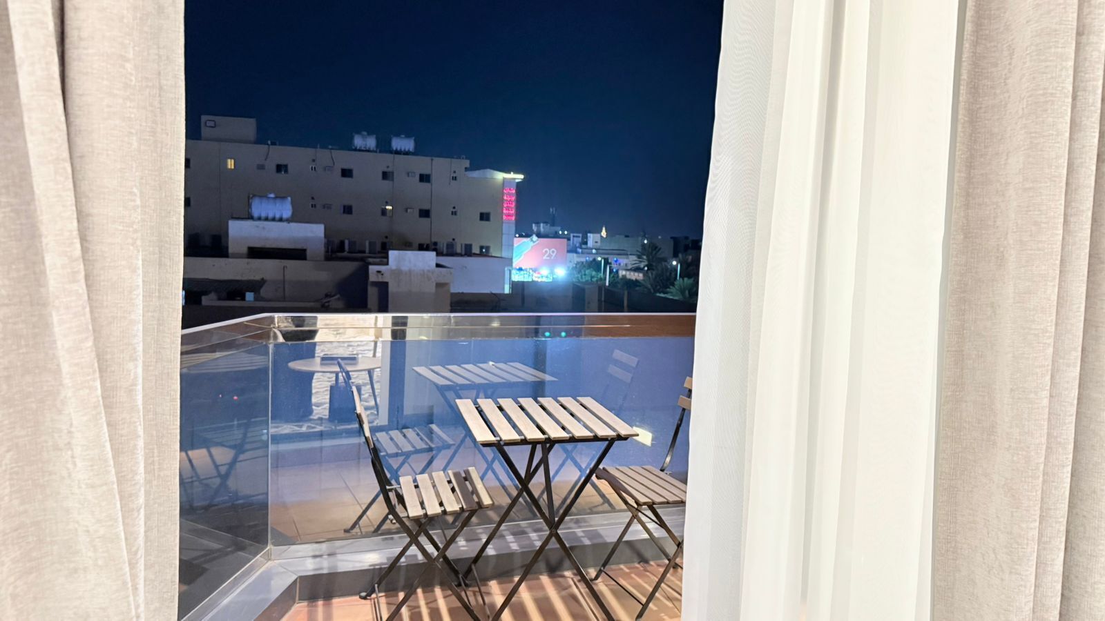 Un balcón acristalado con mobiliario de madera, vistas a un edificio con señalización iluminada y un cielo nocturno despejado.