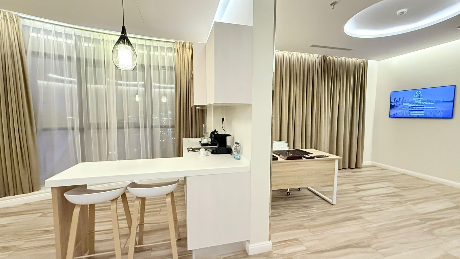 Ein modernes Hotelzimmer mit weißem Schreibtisch, zwei weißen Stühlen, einer kleinen Küchenzeile und einem Fernseher an der Wand.