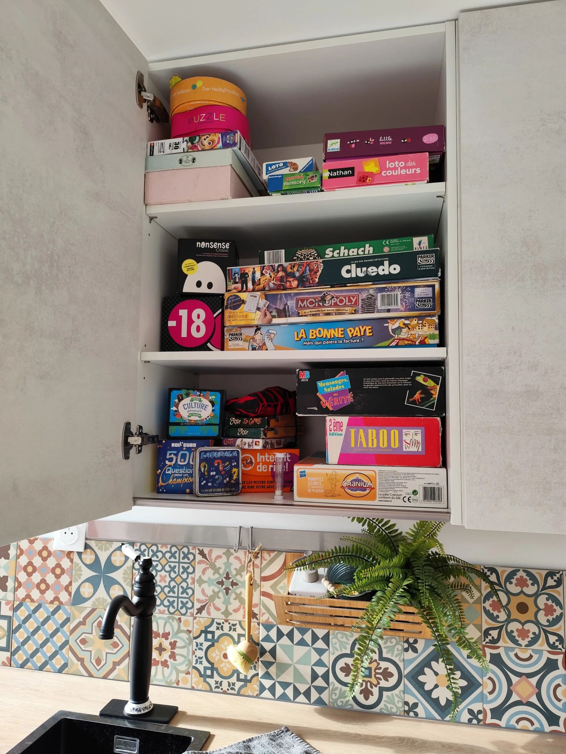 A kitchen with a tiled backsplash, a sink, and a cabinet filled with board games.