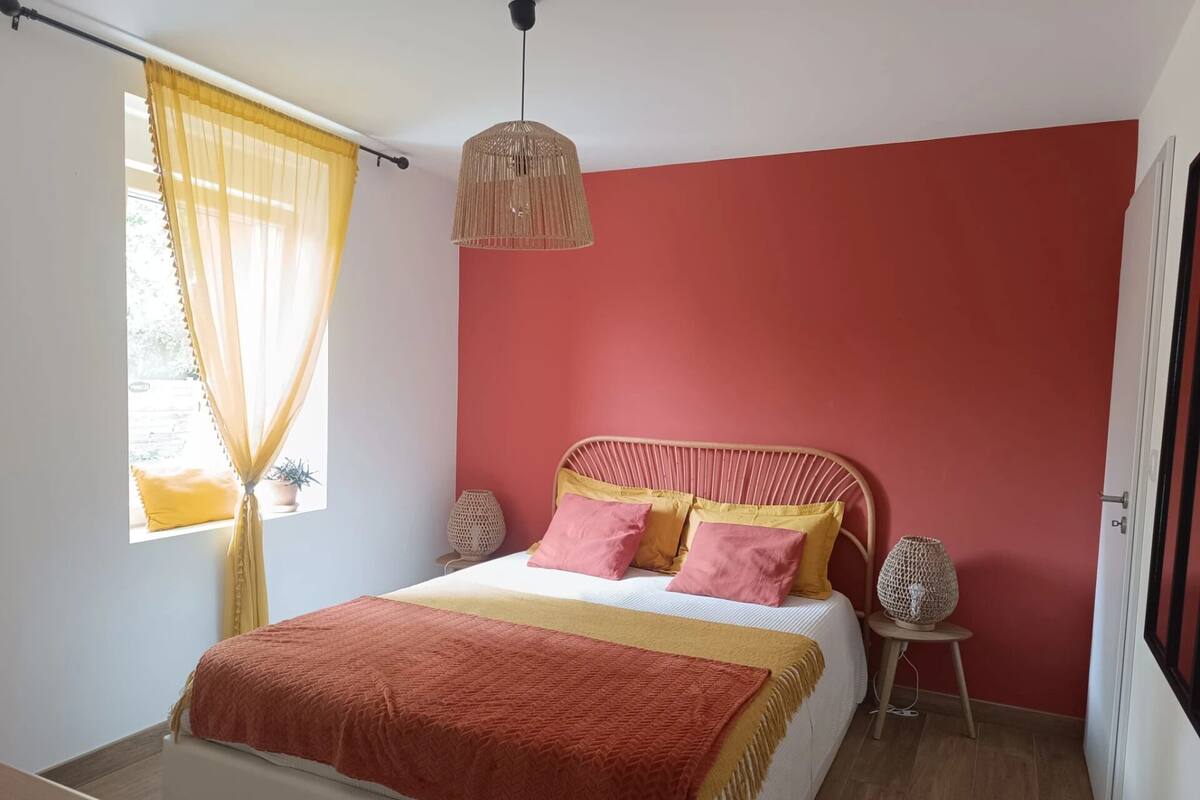 Ein Schlafzimmer mit einem Bett, einem Fenster mit gelben Vorhängen, einer roten Wand, einer Hängeleuchte und einem kleinen Tisch mit einer Lampe.