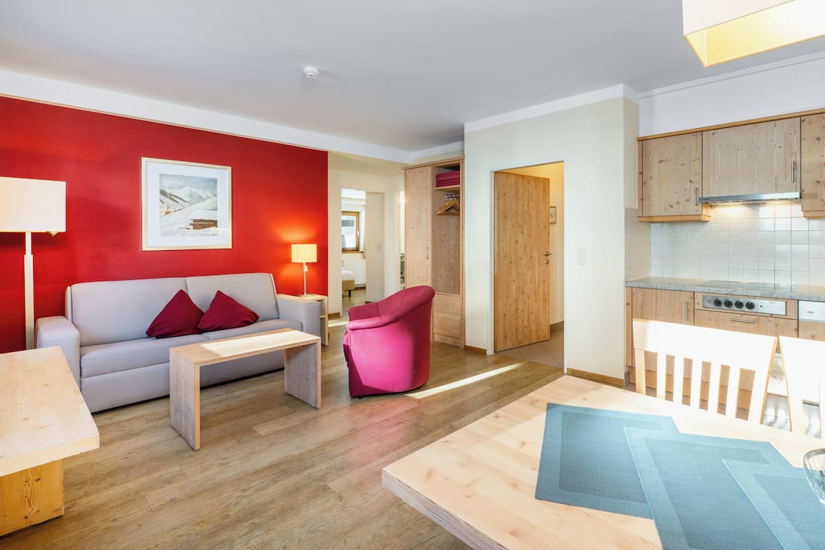 Un salón moderno con una pared de acento roja, un sofá gris, una silla rosa y una mesa de centro de madera. Se ve una cocina con armarios de madera y un horno empotrado al fondo.