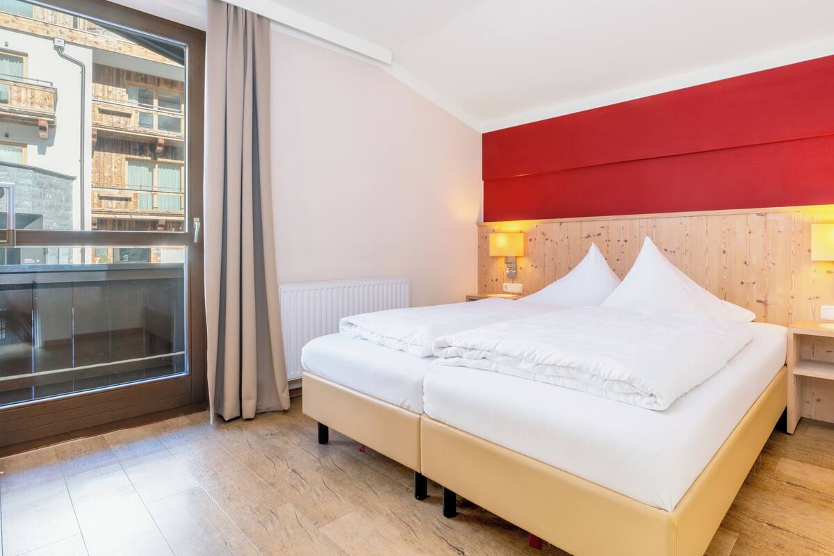 Habitación de hotel con cama, mesita de noche, pared de acento roja, ventana con cortinas y vista a edificios.