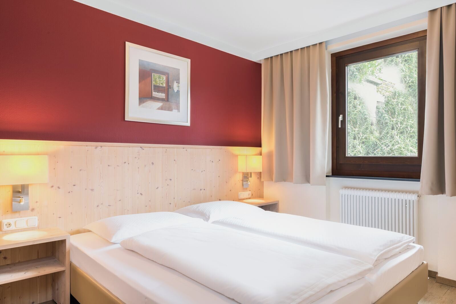 A hotel room with a wooden headboard, two bedside lamps, a window with curtains, and a framed picture on the wall.
