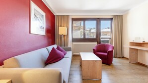 A modern living room with a grey sofa, a red armchair, a wooden coffee table, and a view of a snowy landscape through large windows. - Hapimag Saalbach (Saalbach)