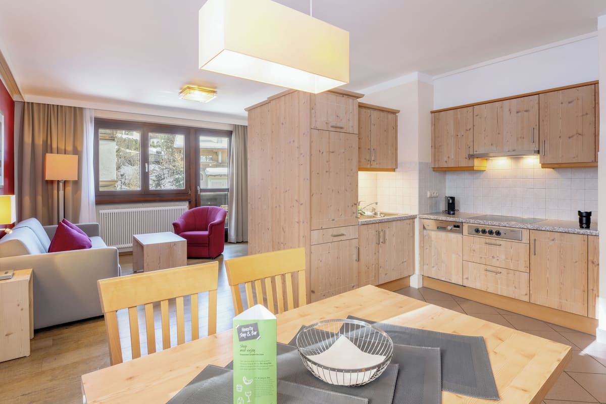 Una habitación de hotel moderna con una mesa de comedor de madera, un sofá, un sillón rojo y una cocina con armarios de madera y una estufa.
