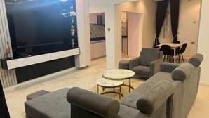 A modern living room with a sectional sofa, a TV, and a dining area with chairs. - Safe Court Apartment (Lekki)