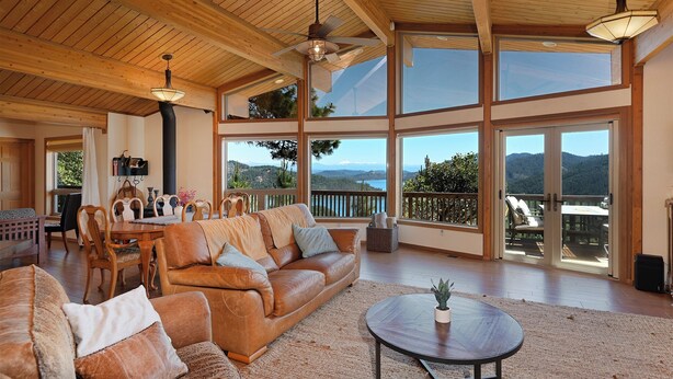 A spacious living room with a large brown leather sofa, wooden dining table, and chairs. There are large windows offering a view of the mountains and a balcony with outdoor seating. - Beautiful Home Above Lake Shasta 30 nights minimum (Lakehead)
