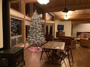 A cozy living room with a wooden ceiling, a large window, a fireplace, a dining table with chairs, and a Christmas tree.