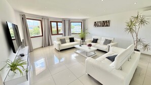 A modern living room with two white sofas, a coffee table, and a flat-screen TV. There are large windows with curtains, a potted plant, and a wall-mounted sign. - Villa with pool in Lecci (Lecci)