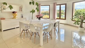 A modern dining area with a white table and chairs, a vase of flowers, and large windows with a view of greenery. - Villa with pool in Lecci (Lecci)