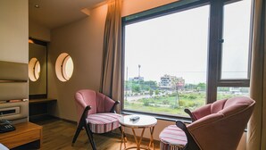 Ein Hotelzimmer mit zwei rosafarbenen Sesseln, einem kleinen runden Tisch, einem Bett, einem Fernseher und einem großen Fenster mit Blick auf Gebäude und Grünflächen.