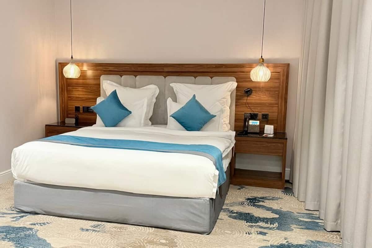 A neatly made bed with a white and blue bedspread, two blue pillows, and a wooden headboard. There are two bedside tables with lamps and a telephone.