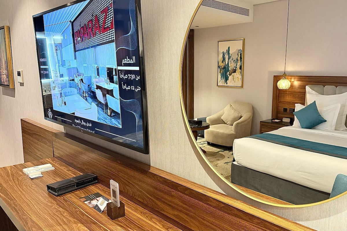 A hotel room with a large TV on the wall, a bed with blue and white bedding, a chair, a bedside table, and a mirror.