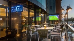 Et udendørs opholdsområde med borde og stole, et stort skærm der viser en sportsbegivenhed, og et skilt for 'Paparazzi Cafe & Restaurant'.