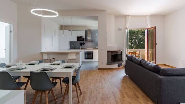 Eine moderne Küche mit weißen Schränken, ein Essbereich mit Tisch für vier Personen und ein graues Sofa in einem Wohnbereich mit Kamin.