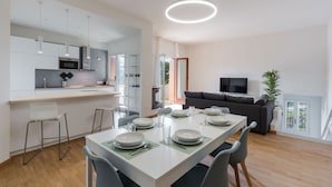 Eine moderne Küche mit weißen Schränken, eine Essecke mit einem langen Tisch, der für vier Personen gedeckt ist, und ein Wohnbereich mit einem dunklen Sofa und einem Fernseher.