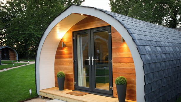 A modern, semi-circular wooden structure with a glass door, two potted plants, and a wooden deck. - Pea Pod Self-Catering glamping in Suffolk (Henley)