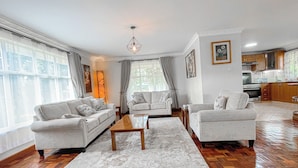 A spacious living room with two sofas, a coffee table, and a rug. There is a kitchen in the background with wooden cabinets and a stove. - Cedar (Athi River)