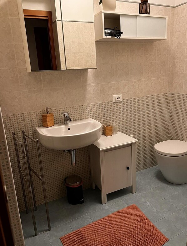 Un baño con lavabo, inodoro y espejo sobre el lavabo. Hay un mueble pequeño encima del lavabo y una repisa con una lámpara.