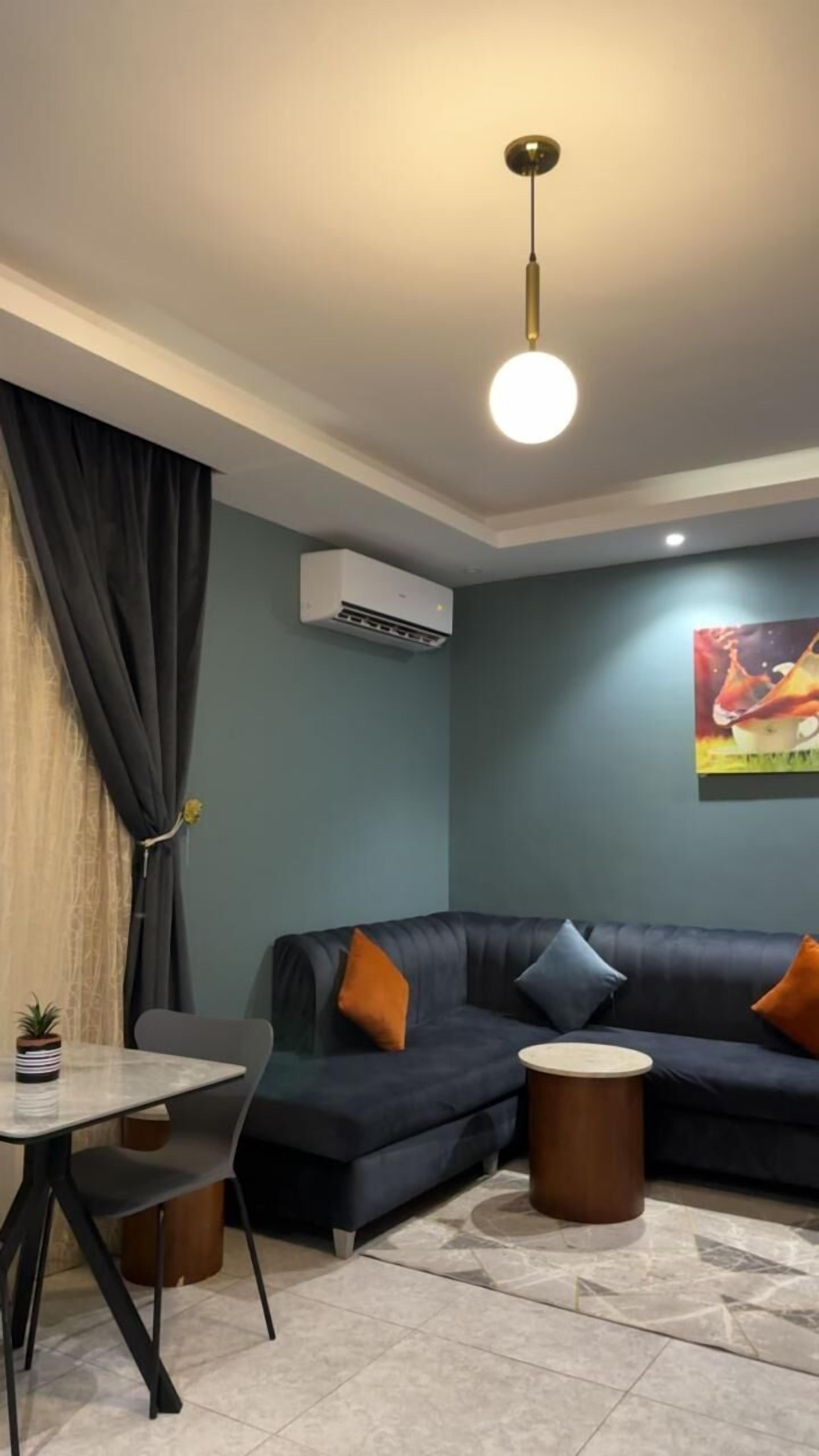 Un moderno salón con un sofá azul oscuro, dos cojines naranja, una mesa redonda de centro, una pequeña planta en maceta y un cuadro en la pared.