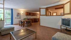 A modern living room with a glass coffee table, a sofa, and a television. There is a dining area with a table and chairs, and a kitchen with wooden cabinets.