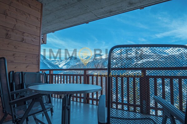 A wooden interior with a round table and chairs, a view of snow-capped mountains, and a metal railing.