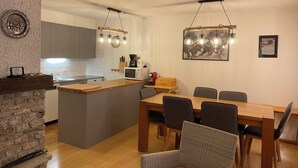 A modern kitchen with a central island, wooden dining table, and chairs. There are hanging light fixtures above the island and a framed picture on the wall.