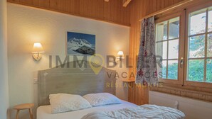 A bedroom with a bed, two wall-mounted lamps, a painting, and a window with curtains. - Located at the entrance to the village, right next to the shuttle bus stop and the supermarket, this (St-Luc)